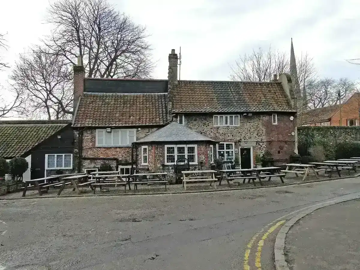 Low flint and brick pub with pantiled roofs and benches outside, the cathedral spire behind