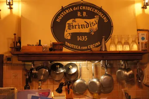 The oval sign inside Al Brindisi reading Hostaria del Chiucchiolino, al Brindisi, 1435, Enoteca con cucina, over a shelf of carafes