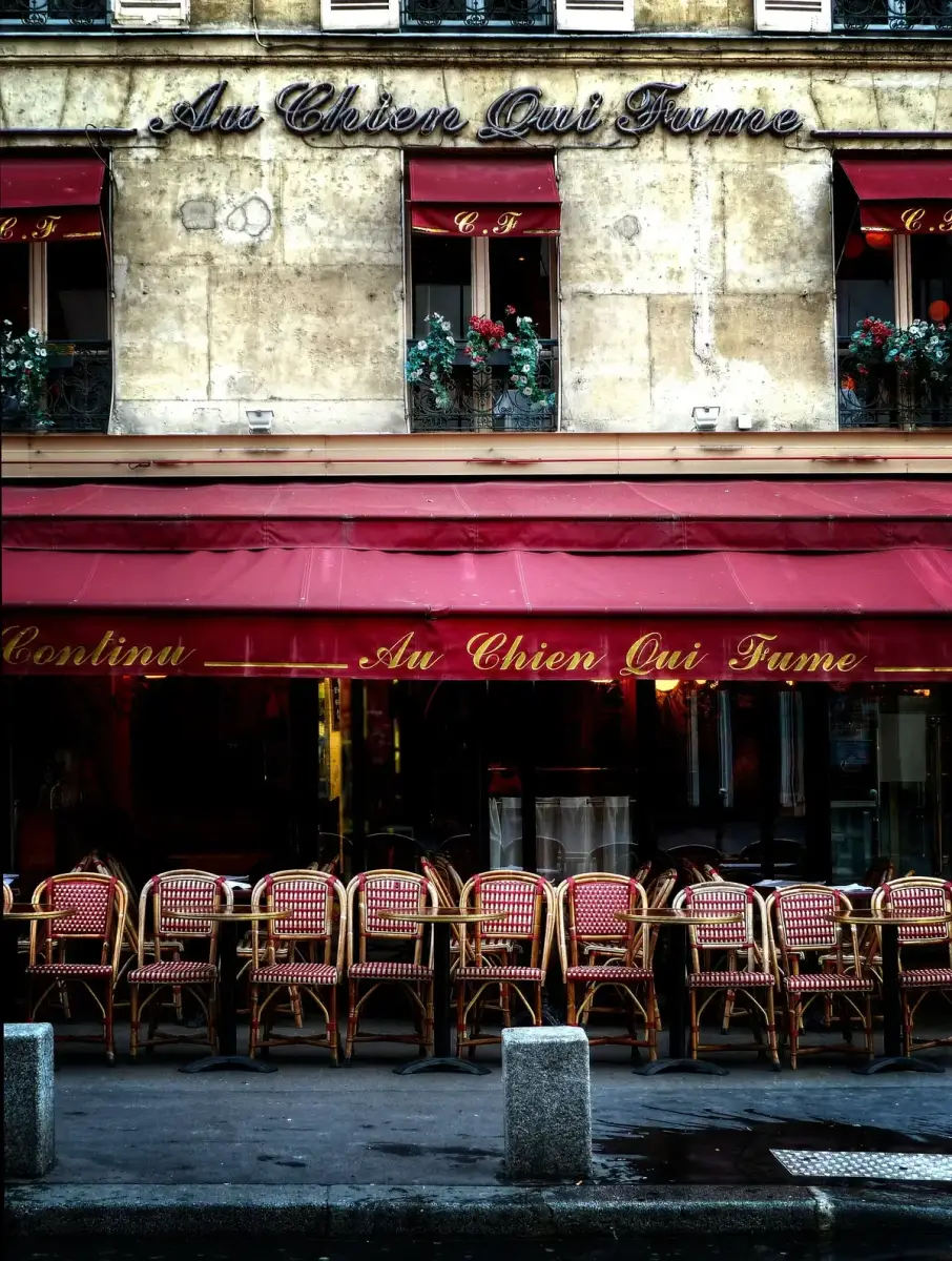 Stone front with Au Chien Qui Fume in neon script, red awnings and rows of café chairs outside