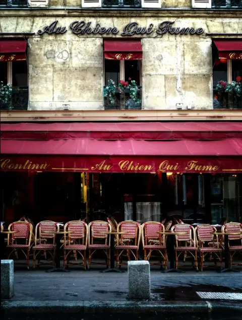 Stone front with Au Chien Qui Fume in neon script, red awnings and rows of café chairs outside