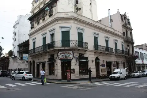Pale corner building with iron balconies and green shutters above a painted bar sign
