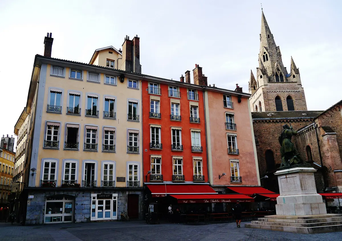 Tall painted houses on place Saint-André, Grenoble, with the red awnings of La Table Ronde along the ground floor and the collegiate church spire at right
