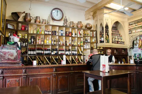Long wooden counter under shelves of bottles, copper jars and a clock, a customer served at the bar