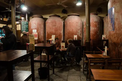 Back room with tall clay wine jars ranged along the wall and drinkers at plain wooden tables