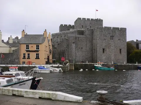 Apricot pub lettered The Castle Arms beside the grey walls of Castle Rushen, seen over the harbour