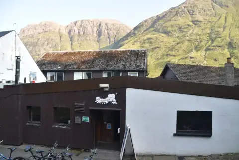 Low timber-clad bar entrance signed Boots Bar, under the steep green flank of the Glencoe ridge