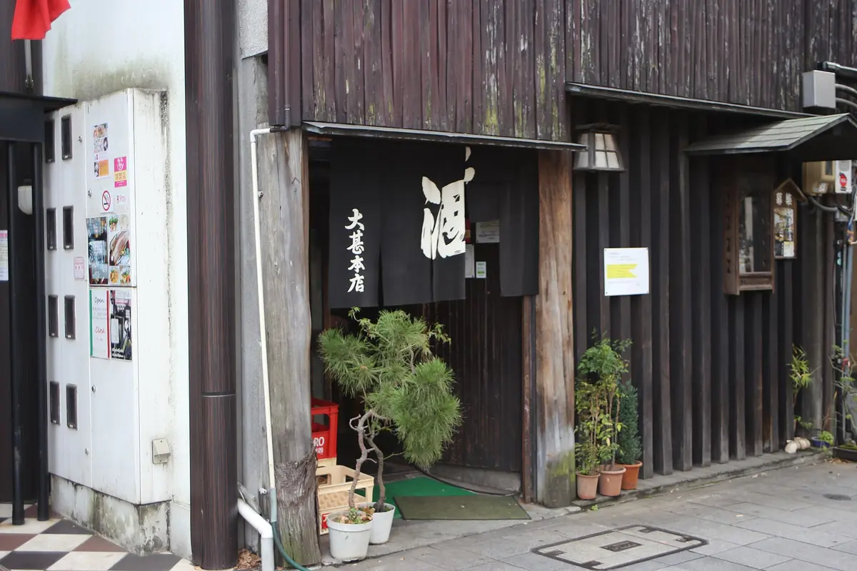 Dark timber shopfront with a black noren reading sake and Daijin Honten, a potted pine beside the door