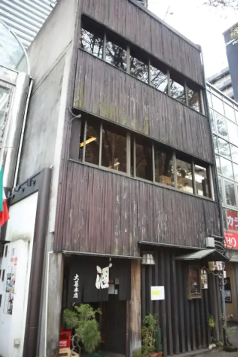 Three-storey street front boarded in weathered dark timber, the izakaya’s noren hanging at the door below
