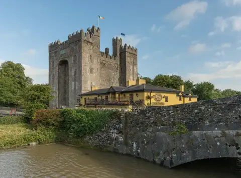 Yellow-washed pub with a slate roof and flower boxes below the grey keep of Bunratty Castle