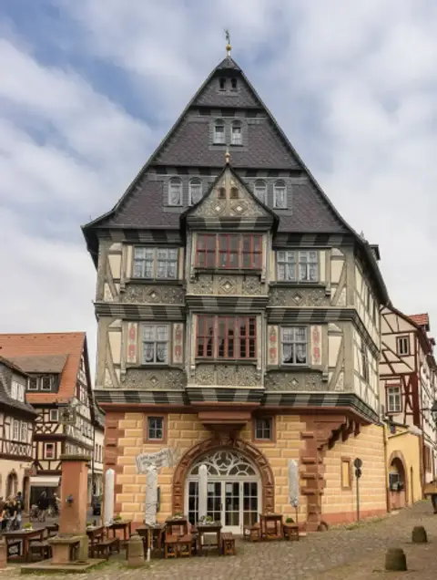Front of the same half-timbered inn with an arched doorway and benches on the cobbled square