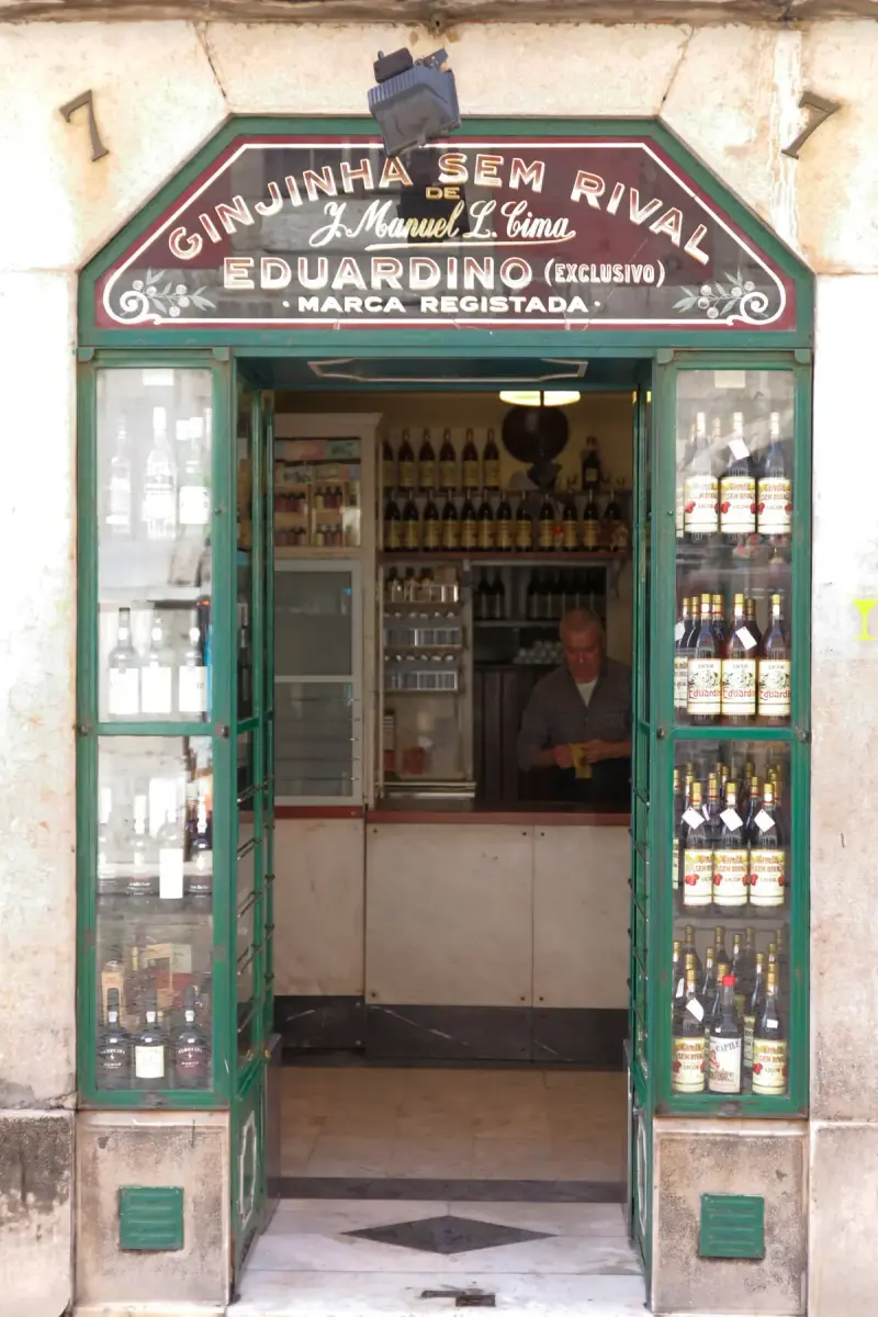 Green-framed doorway of Ginjinha Sem Rival at number 7, its arched sign over the door and cabinets of ginja bottles either side of the counter