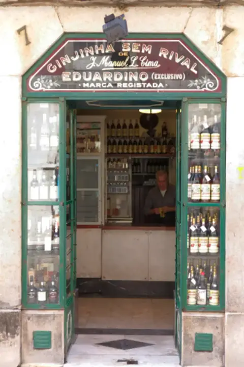 Green-framed doorway of Ginjinha Sem Rival at number 7, its arched sign over the door and cabinets of ginja bottles either side of the counter