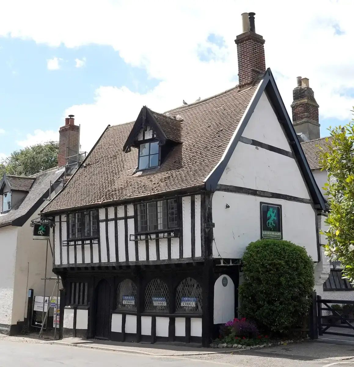 Jettied timber-framed inn with a dormer and a green dragon sign, brick stacks above