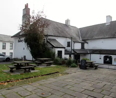 Whitewashed pub with slate roofs around a flagged yard of picnic tables, its name on the gable