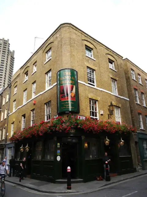 Yellow-brick corner pub with a curved green shopfront, lanterns and a band of red flower boxes