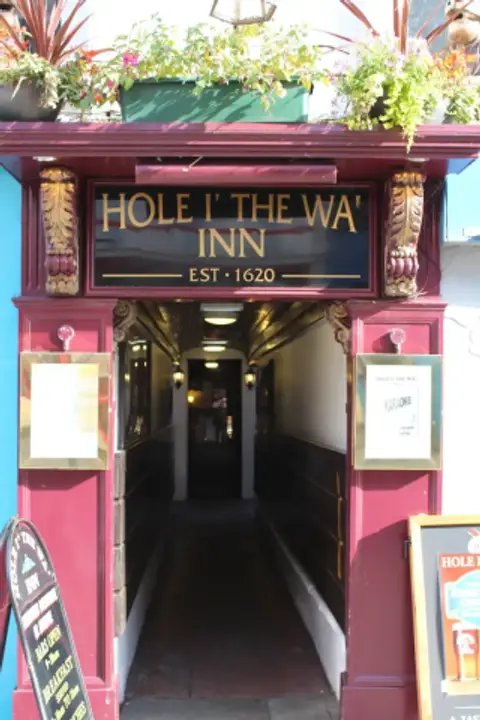 A narrow pub entrance painted deep red, its fascia lettered in gold: Hole i the Wa Inn, est 1620, above a passage running back from the street
