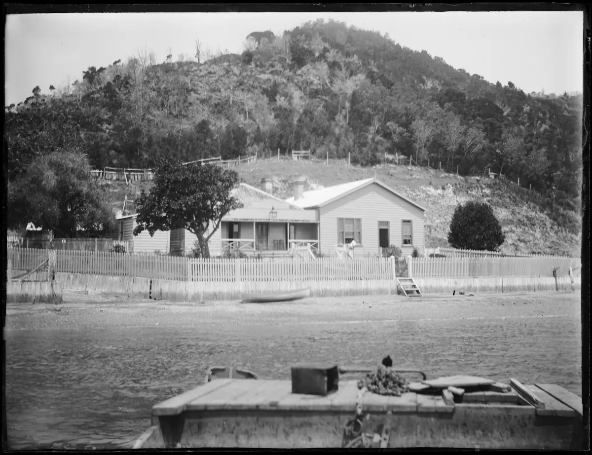 Black-and-white photograph of a weatherboard hotel behind a picket fence on the Horeke waterfront, bush-covered hill behind and a punt in front