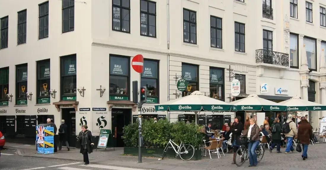 Cream corner building with green awnings and dark green painted lettering, tables on the pavement