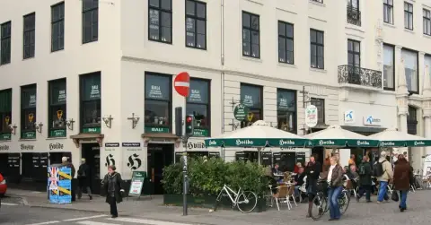 Cream corner building with green awnings and dark green painted lettering, tables on the pavement