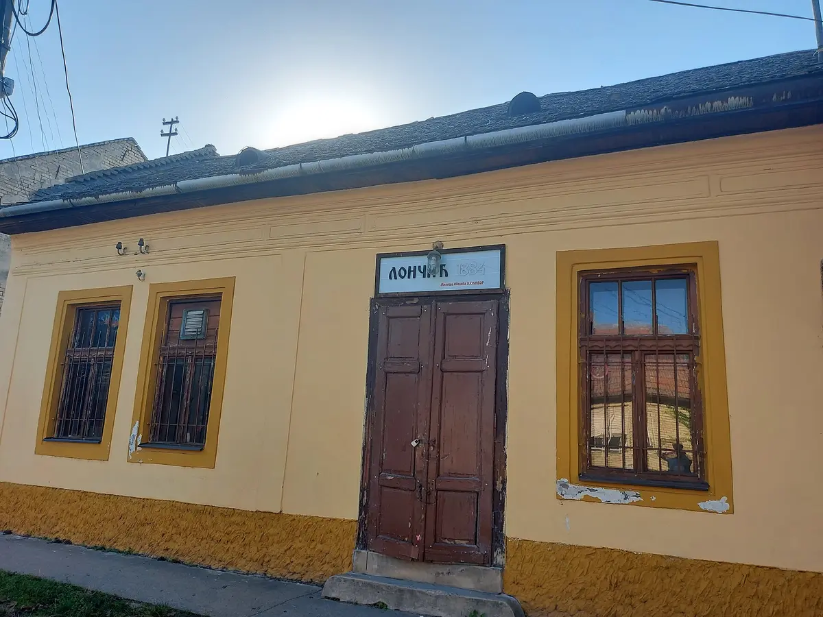 Single-storey ochre house with barred windows and a Лончић 1884 board over brown double doors