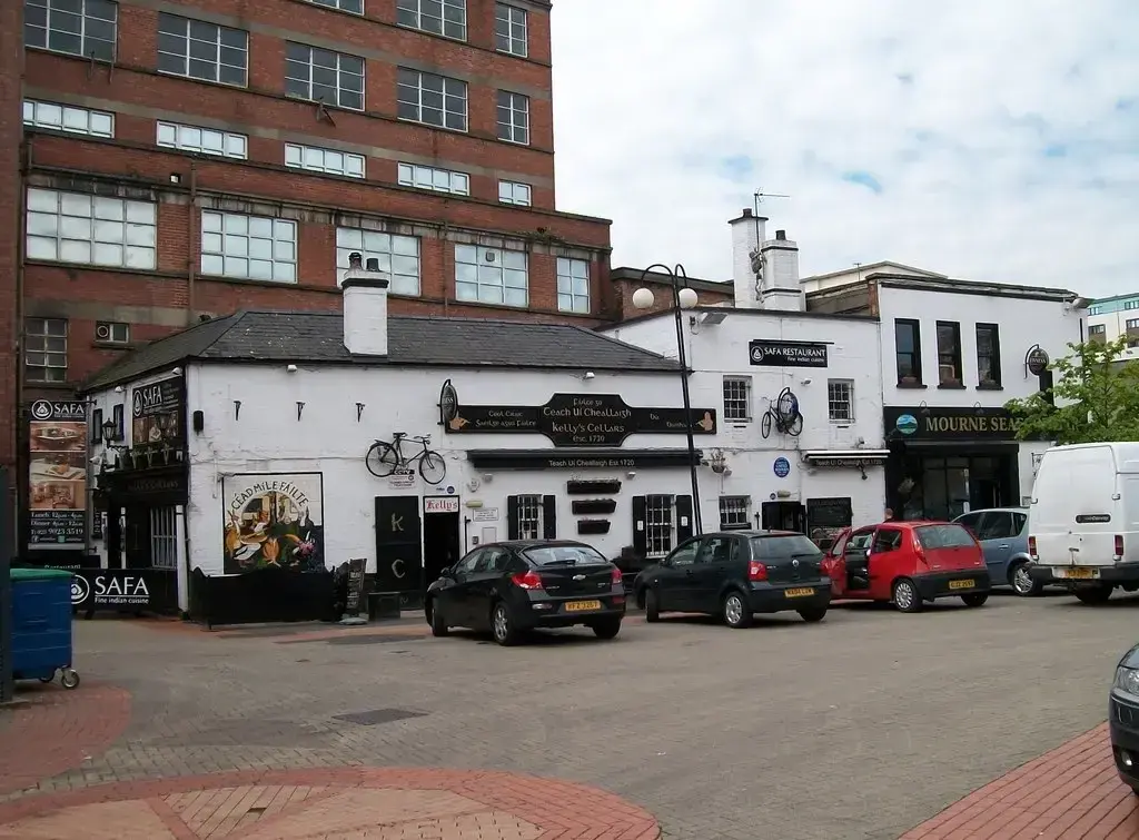 Low whitewashed pub with a slate roof and black fascia, hemmed in by taller modern buildings