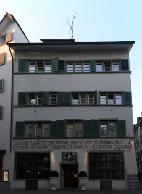 Four-storey Zürich house at dusk, lit restaurant windows below a red painted inscription