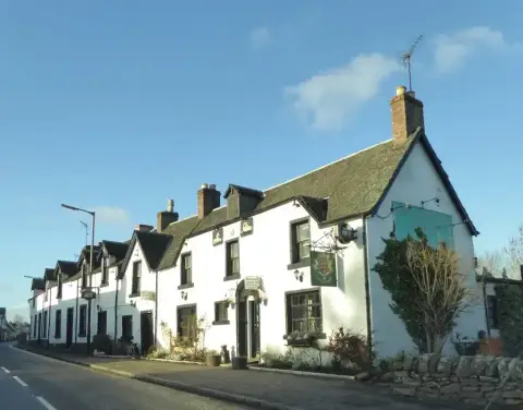 Long white harled inn with dormer windows and a hanging sign, running down a village street
