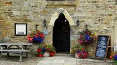 Pointed stone arch doorway in a rubble-stone wall, flanked by hanging baskets and a chalkboard