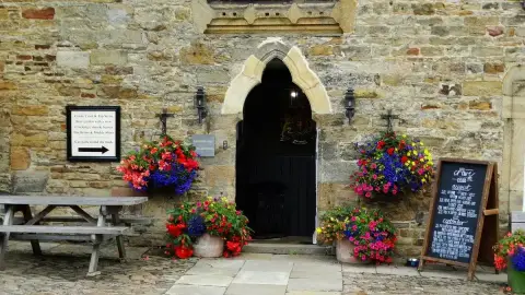 Pointed stone arch doorway in a rubble-stone wall, flanked by hanging baskets and a chalkboard