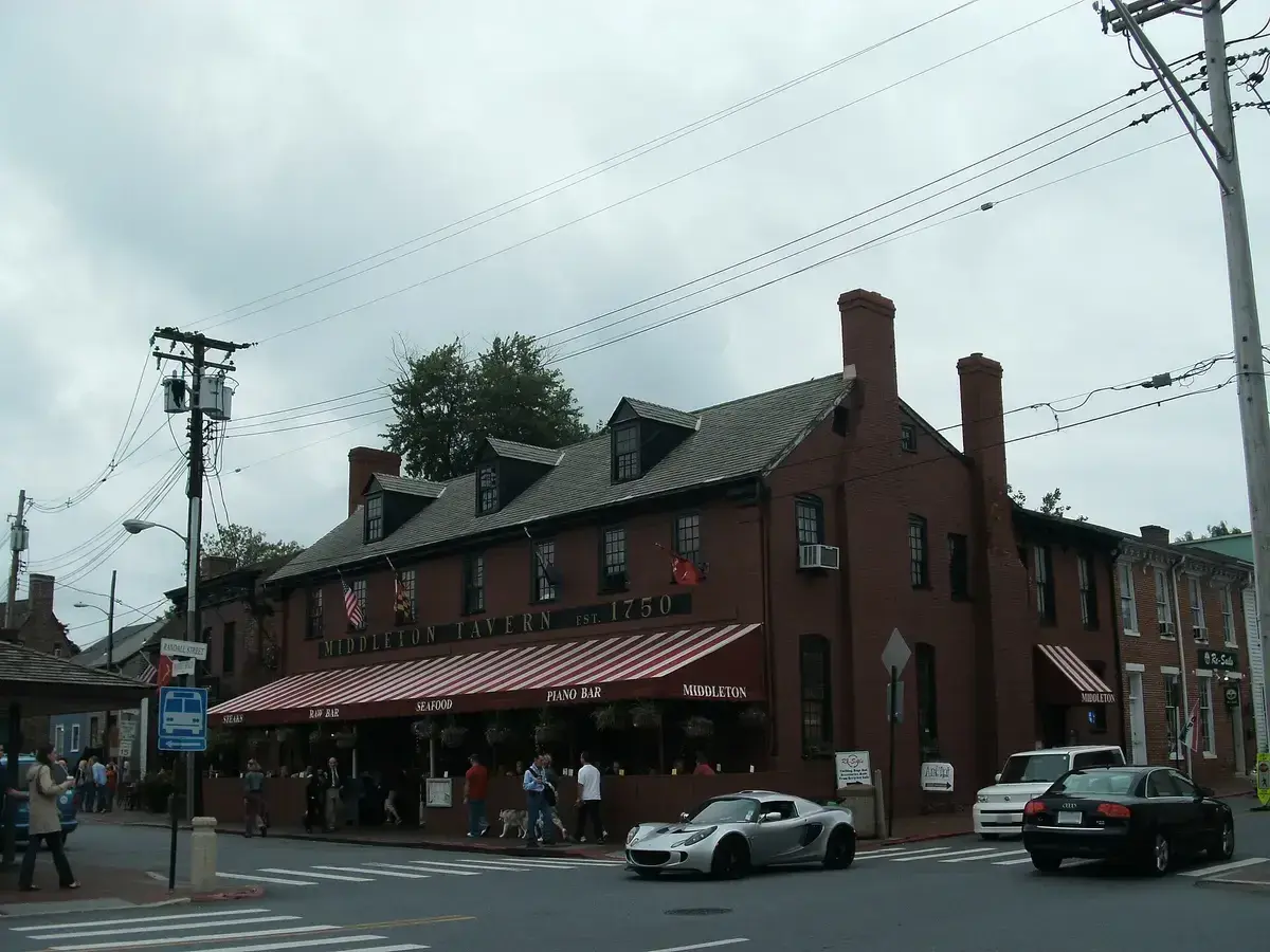 Dark red brick tavern with dormer windows, tall chimneys and a striped awning over the pavement