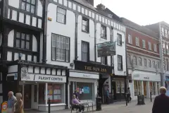 White-rendered galleried inn front between high street shops, gold lettering over the carriage arch