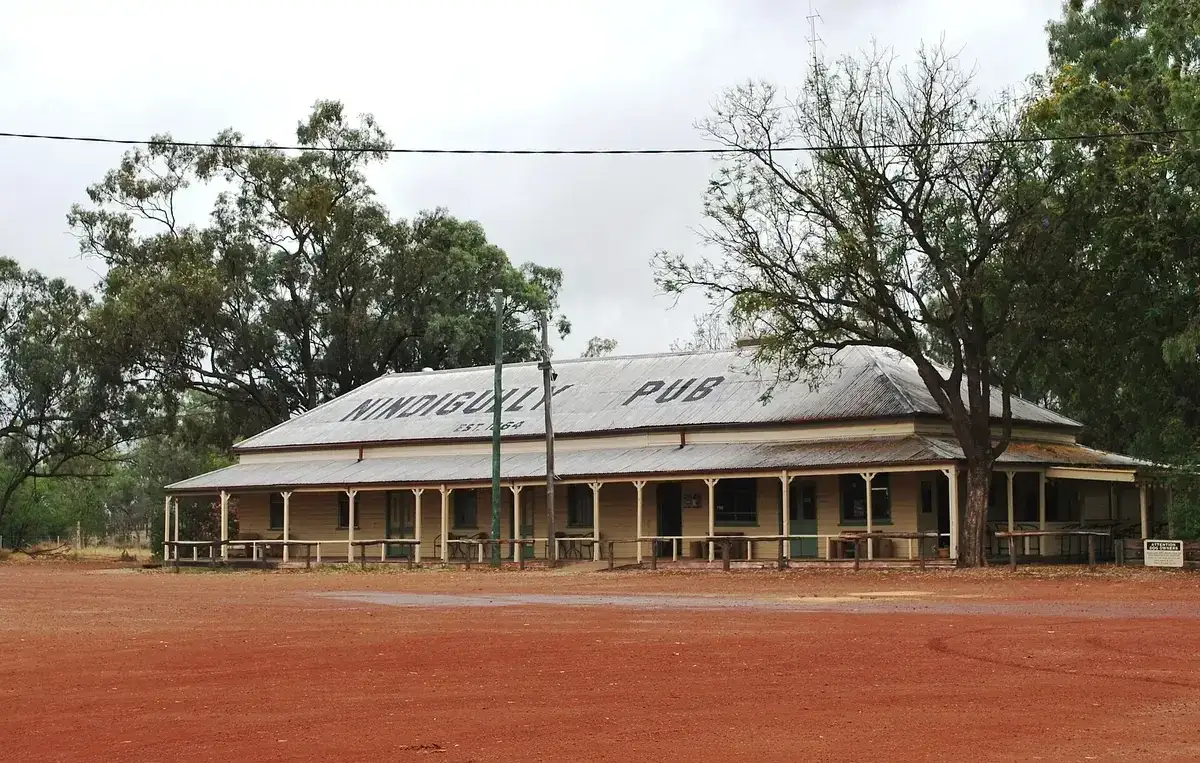 Long timber pub with a corrugated iron roof lettered with its name, verandah facing red earth