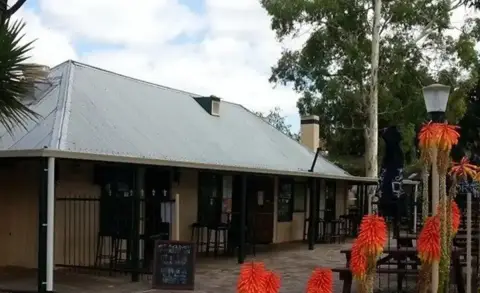 Low slab-and-timber inn with a corrugated iron roof and wide verandah, red-hot pokers in front