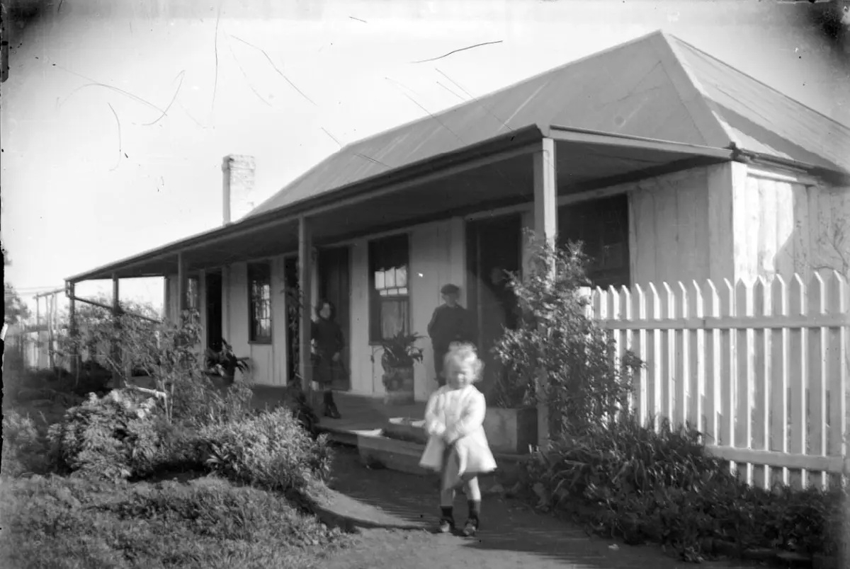 The slab-walled inn with its long verandah in 1910, a small child and two adults posed by the picket fence