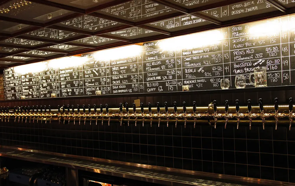 Bar counter with dozens of brass taps in a row under a chalkboard list of beers