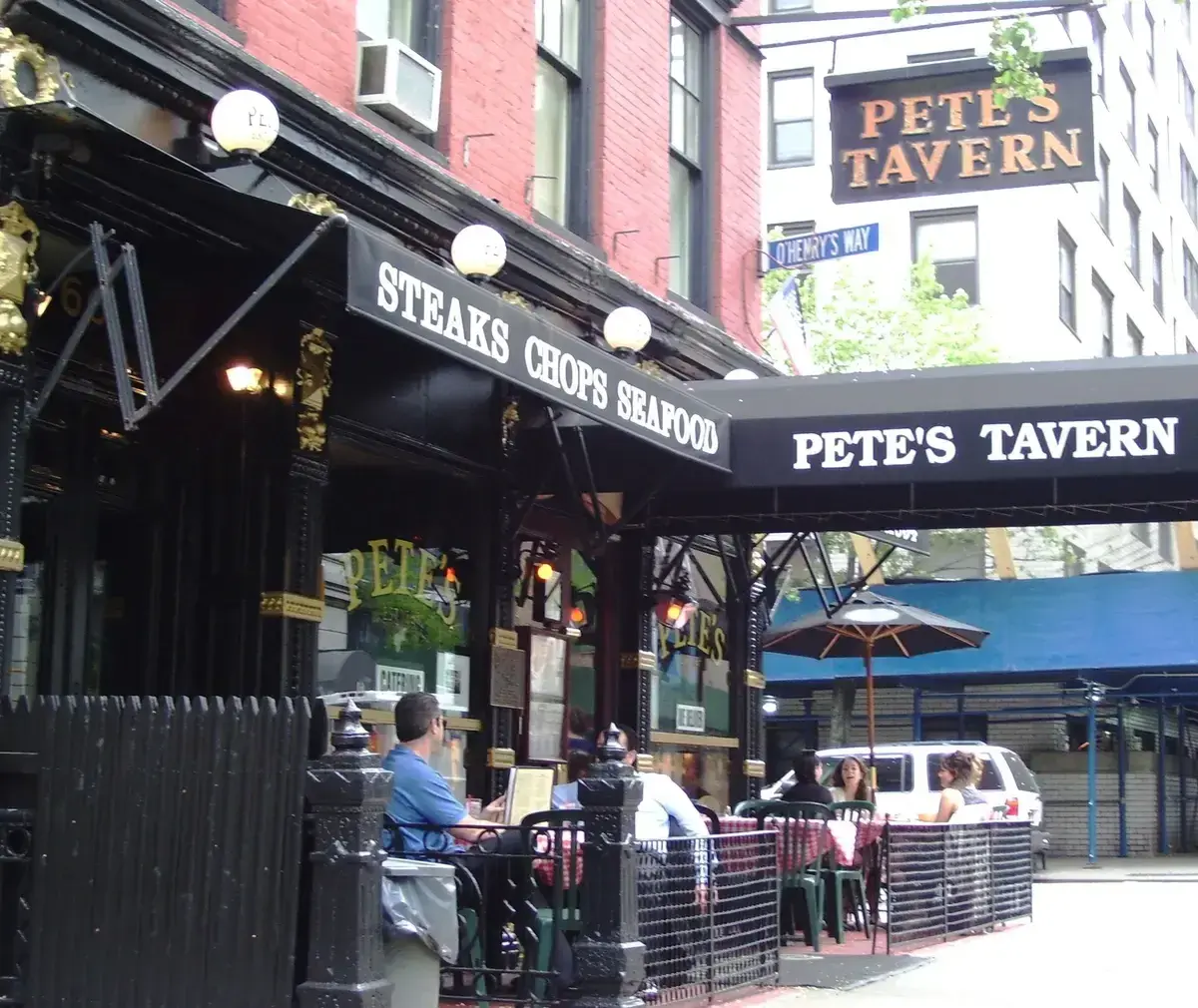 Black cast-iron and glass tavern front with painted awnings and a corner sign, tables outside