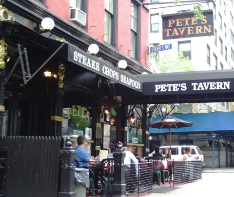 Black cast-iron and glass tavern front with painted awnings and a corner sign, tables outside