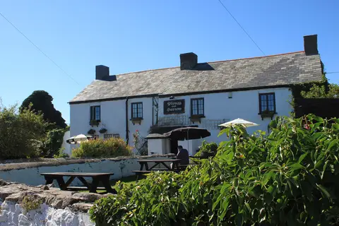 A long whitewashed farmhouse under a slate roof, its hanging sign lettered Plough and Harrow, with benches on the grass in front