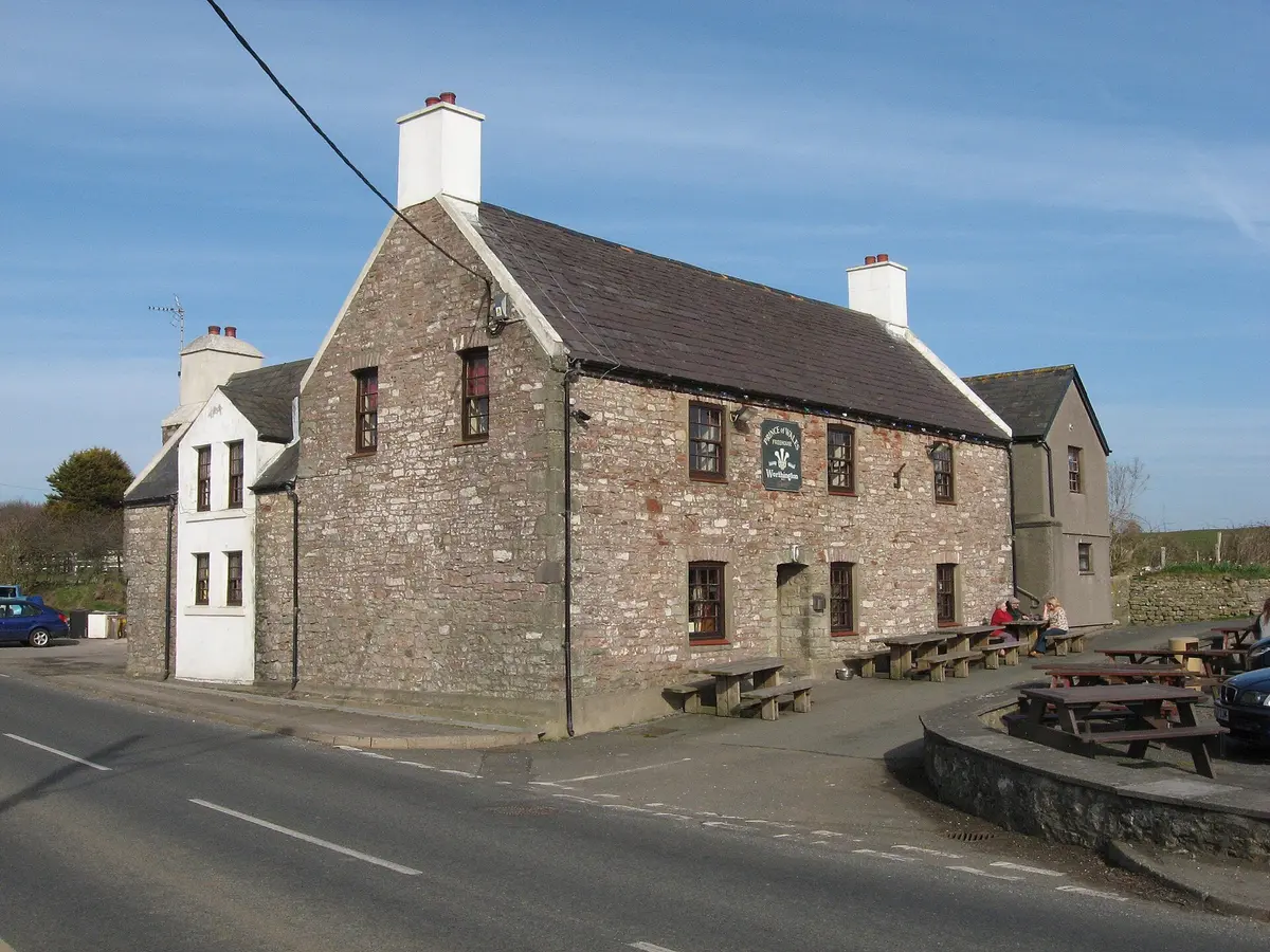 Two-storey rubble-stone pub with white chimneys on a road corner, benches and a green name sign