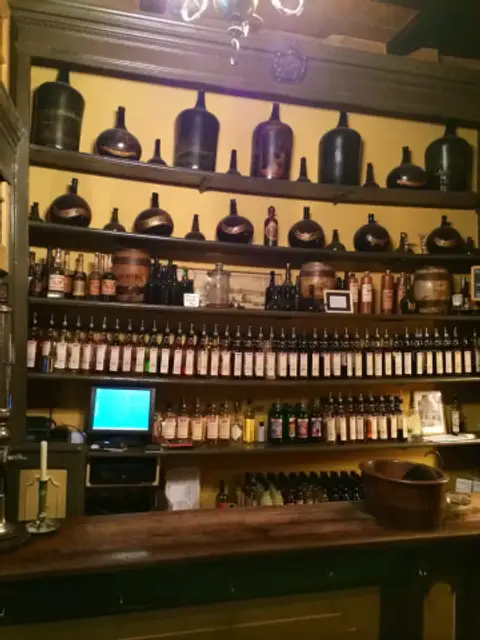 Wooden shelves behind a bar filled with rows of labelled liqueur bottles and old stoneware jars