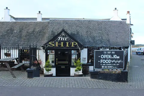 A low white harbour pub under a slate roof, THE SHIP in gold on the porch and a chalked board beside the door