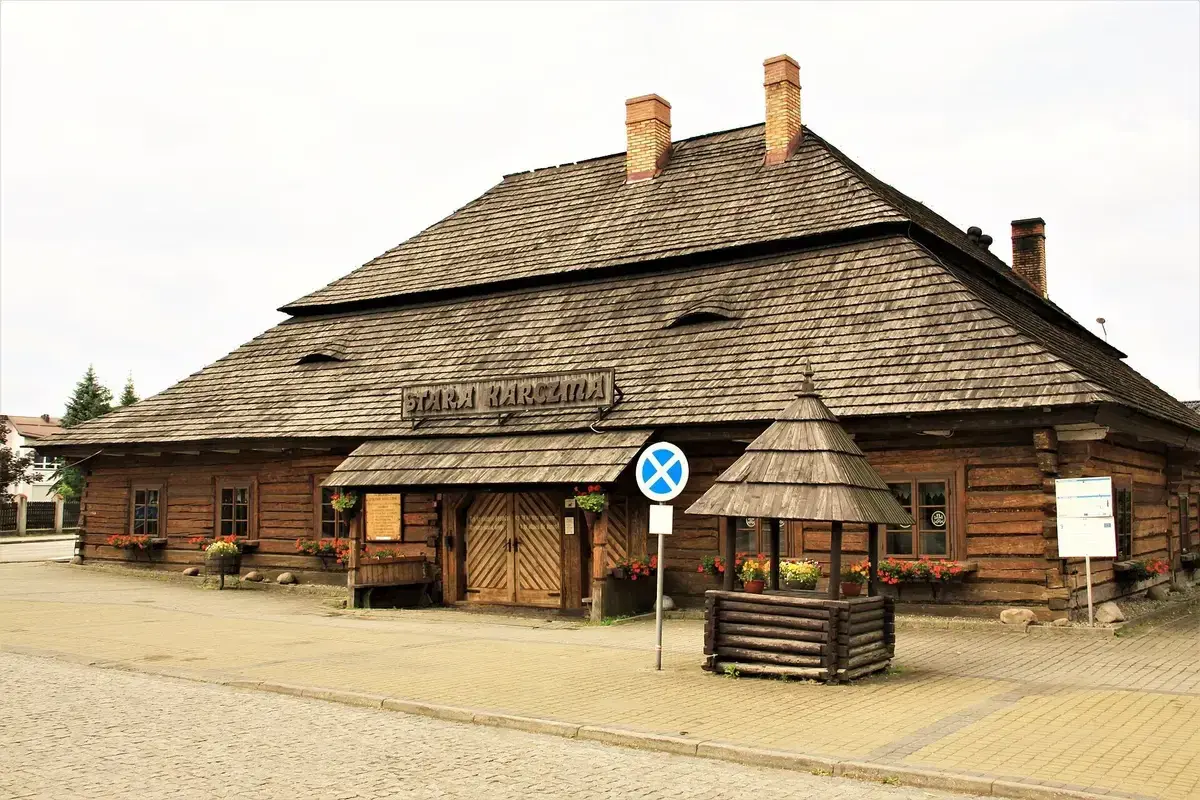 Log-built inn with a huge shingled hipped roof, brick chimneys and flower boxes, beside a well