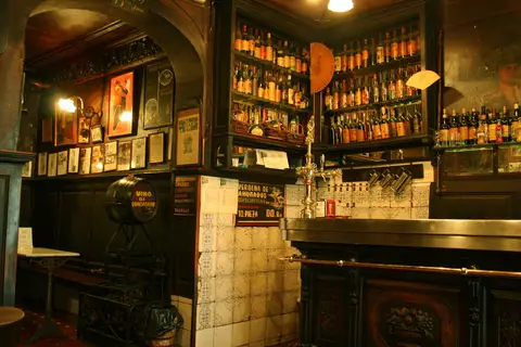 Long counter with a brass vermouth tap, tiled piers and shelves of bottles under framed prints
