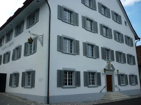 Corner of a white tavern, a gilded crown on a wrought-iron bracket and TAVERNE ZUR KRONE on the gable