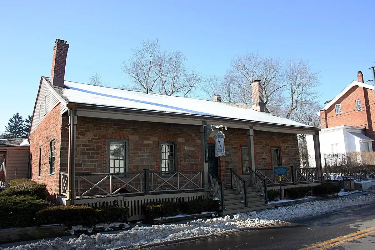 Long single-storey sandstone tavern with a shingled roof, brick chimneys and a porch rail
