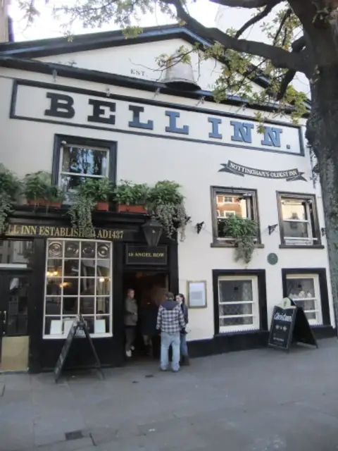 Cream-painted inn front with large black lettering, a gilded bell on the parapet and window plants