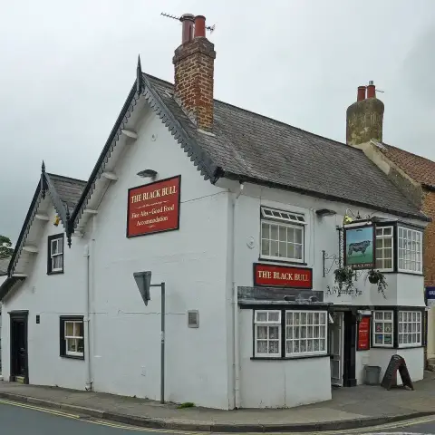 Whitewashed corner inn with bargeboarded gables, red name boards and a bull sign in a wrought-iron bracket