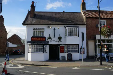 Whitewashed inn on a street corner under blue sky, bay windows and a board reading A 13th Century Inn
