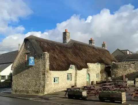 Thick-walled limestone inn under a deep thatched roof, with stone chimneys and green windows
