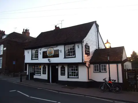White weatherboarded corner pub at dusk, lit lamp and a painted coat-of-arms board on the wall